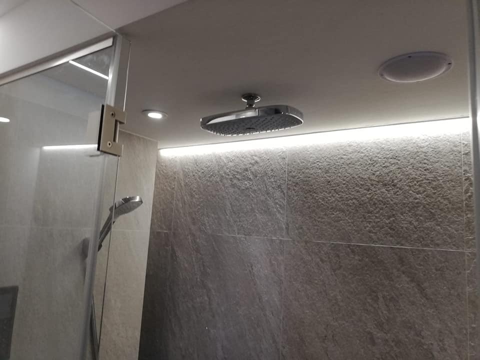 Nowoczesna kabina prysznicowa z deszczownicą, oświetleniem LED i szklanymi drzwiami z metalowymi zawiasami, wykończona płytkami imitującymi kamień.