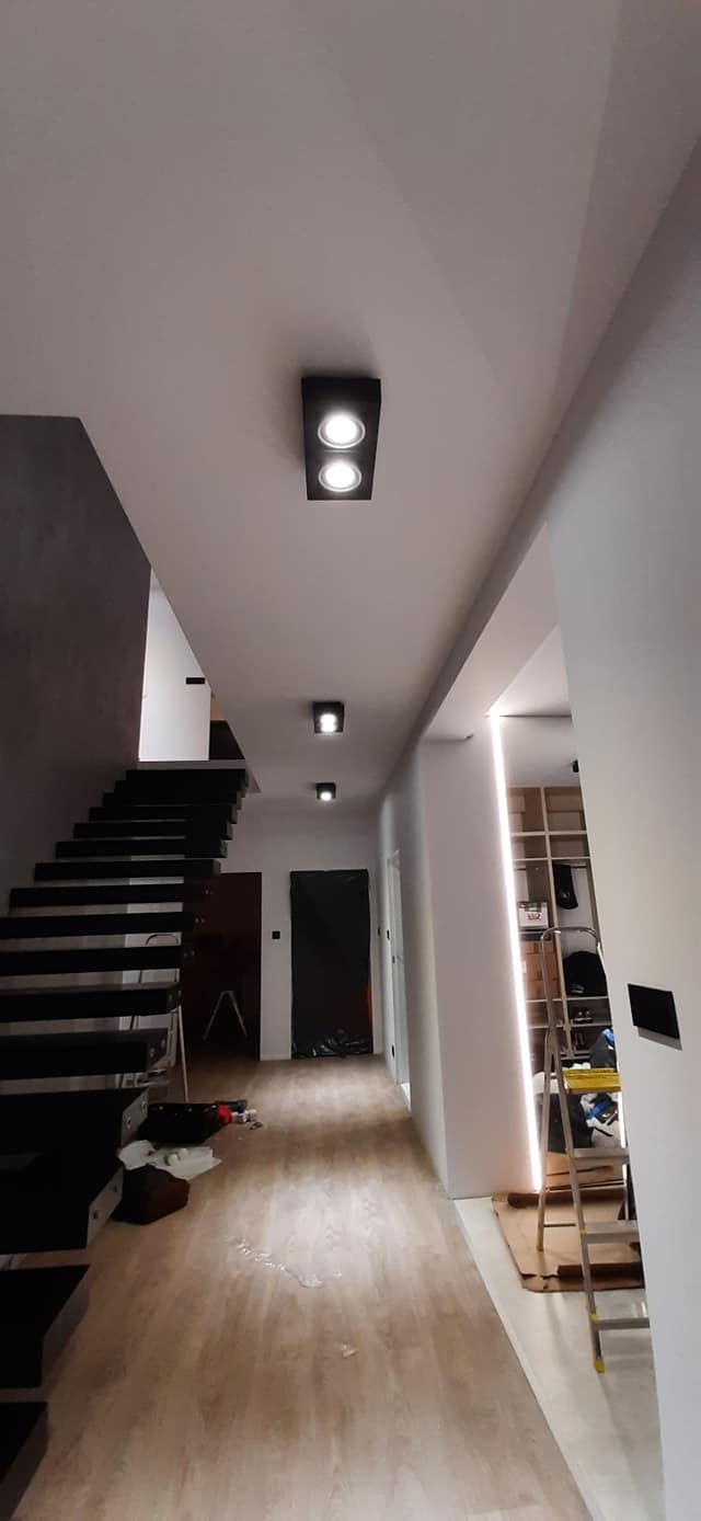 Nowoczesne oświetlenie LED w korytarzu z drewnianą podłogą i schodami, widoczne czarne oprawy z dwoma punktami świetlnymi oraz liniowe oświetlenie wnękowe, częściowo zakryte folią drzwi i drabina...