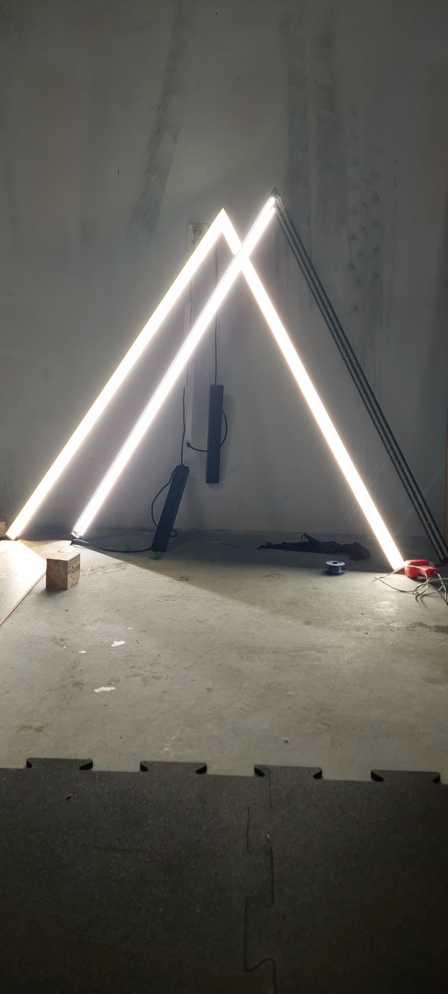 Trójkątna konstrukcja z jasnych, liniowych lamp LED oparta o ścianę w surowym wnętrzu, widoczne przewody i elementy montażowe, podłoga z szarego betonu i czarnych mat.