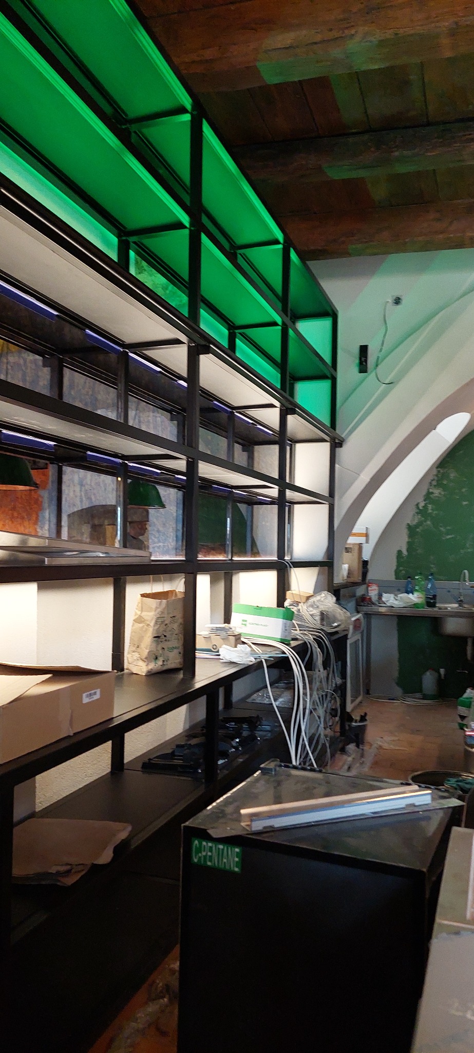 Wnętrze z metalowym regałem podświetlonym na zielono, widoczne kable, kartony i elementy wyposażenia kuchni w tle.