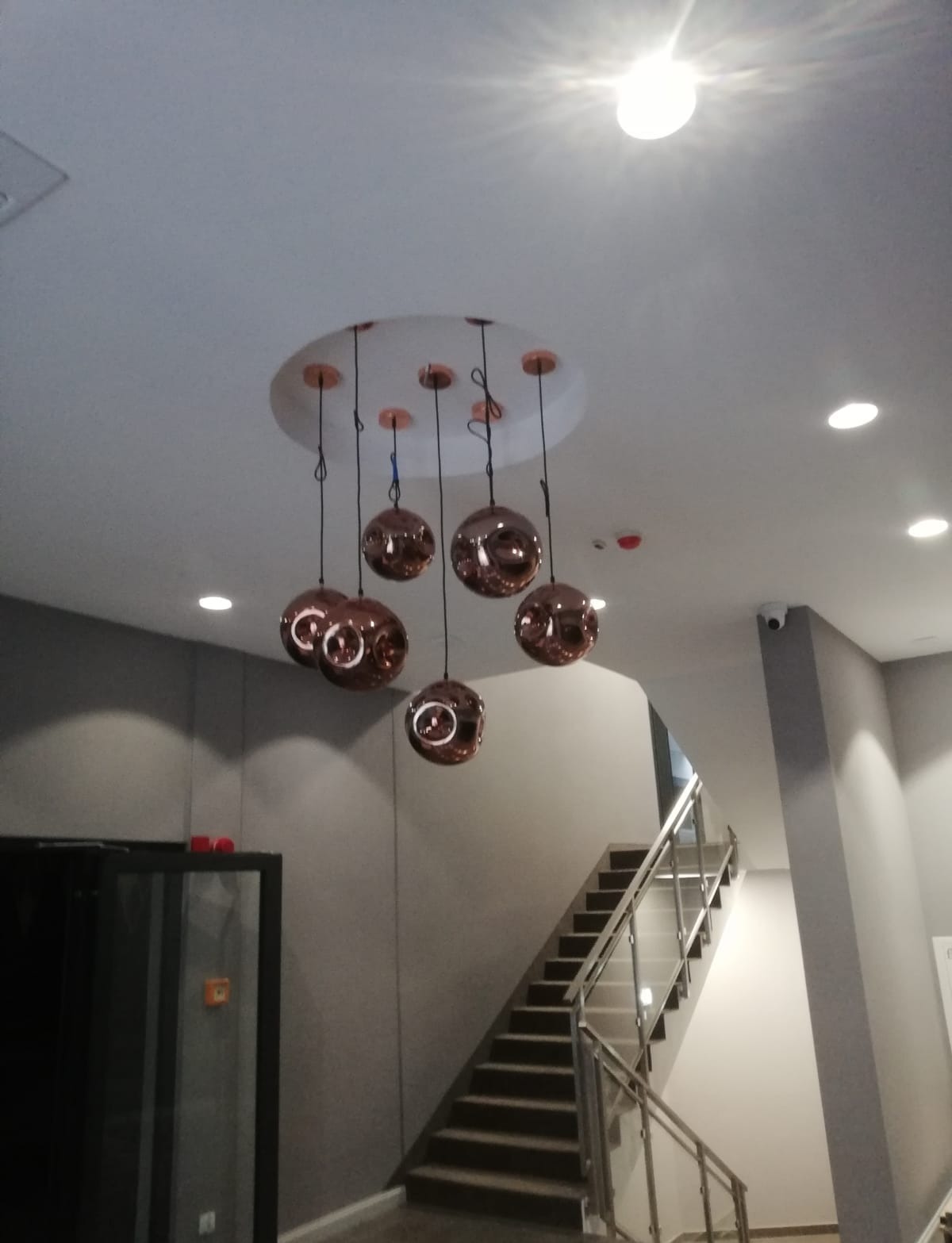 Sufit z zamontowaną dekoracyjną lampą wiszącą z siedmioma kulistymi kloszami w kolorze miedzi, widoczne schody z metalową barierką w tle.