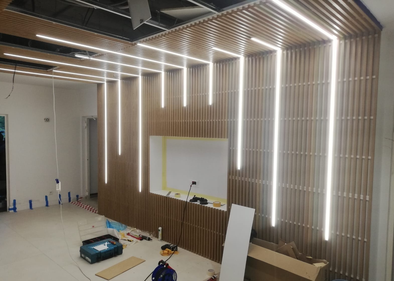 Montaż oświetlenia LED w drewnianej zabudowie sufitu i ścian, widoczne narzędzia i materiały budowlane, surowy stan wykończenia wnętrza.