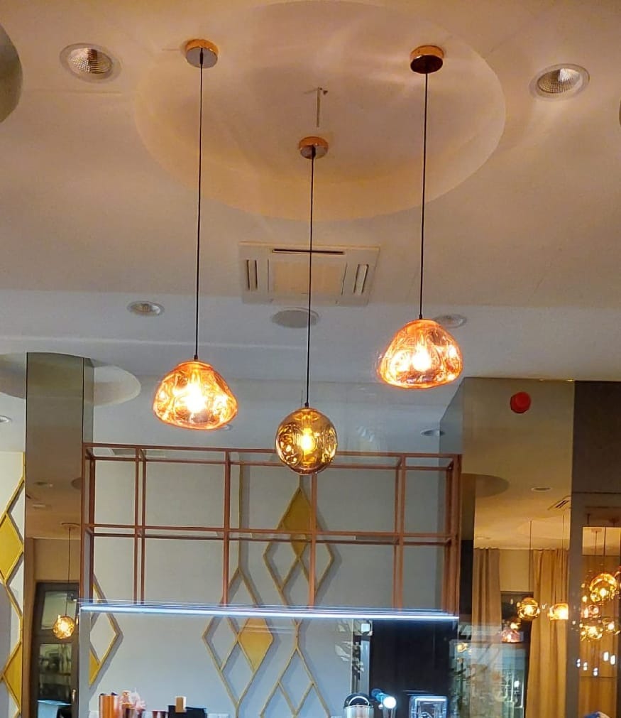 Trzy designerskie lampy wiszące z pomarańczowego szkła o nieregularnym kształcie, zamontowane na tle sufitu z okrągłym wgłębieniem i kratownicy z metalowych profili.