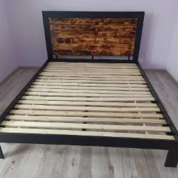 Metalowa rama łóżka z drewnianym wezgłowiem o wypalanej fakturze, widoczne drewniane listwy stelaża.