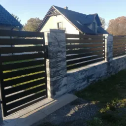 Nowoczesna brama wjazdowa i ogrodzenie z poziomych, ciemnych przęseł metalowych, osadzone na betonowych słupkach i podmurówce z imitacją kamienia, na tle domu jednorodzinnego w słoneczny dzień.