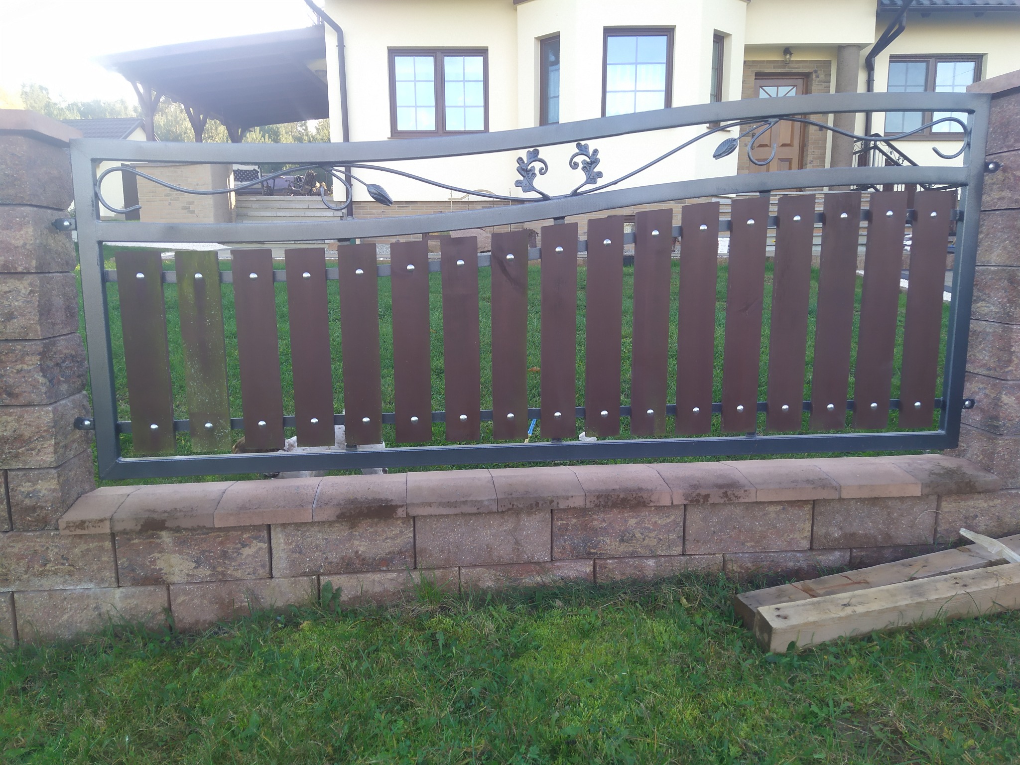 Fragment ogrodzenia: metalowa rama z drewnianymi panelami, osadzona na murku z bloczków, z tłem budynku mieszkalnego i zielonym trawnikiem.