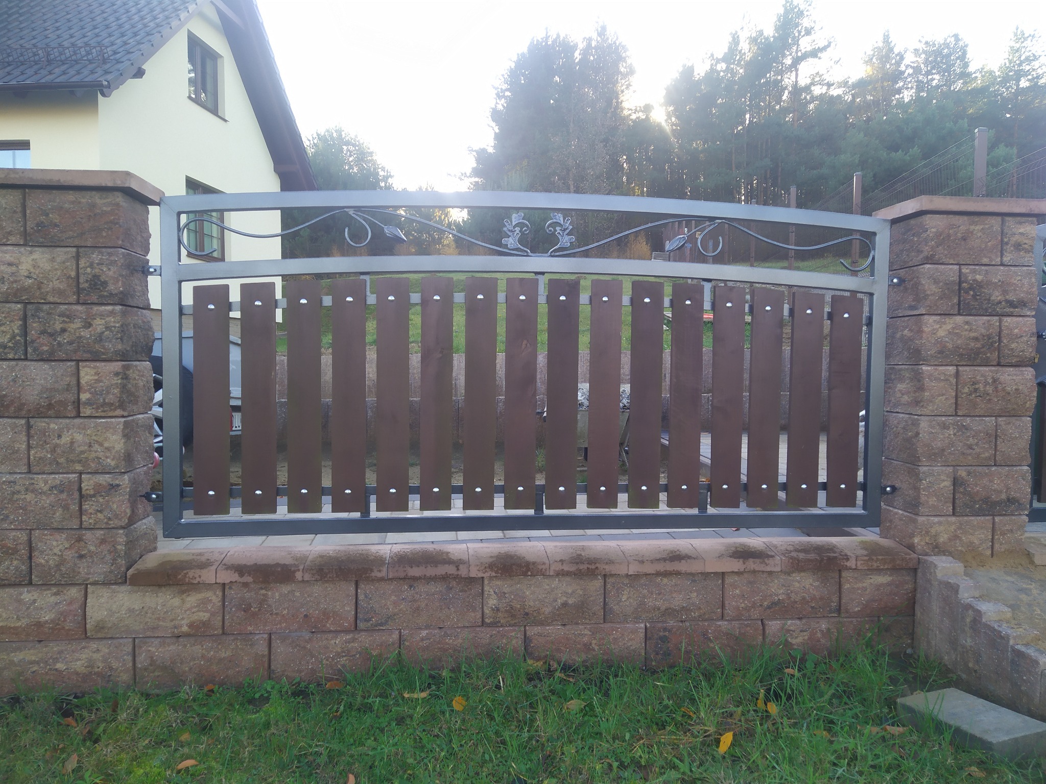 Nowoczesna brama wjazdowa z brązowymi, pionowymi deskami i metalową ramą z ozdobnymi elementami, osadzona na murowanych słupkach z brązowego kamienia.