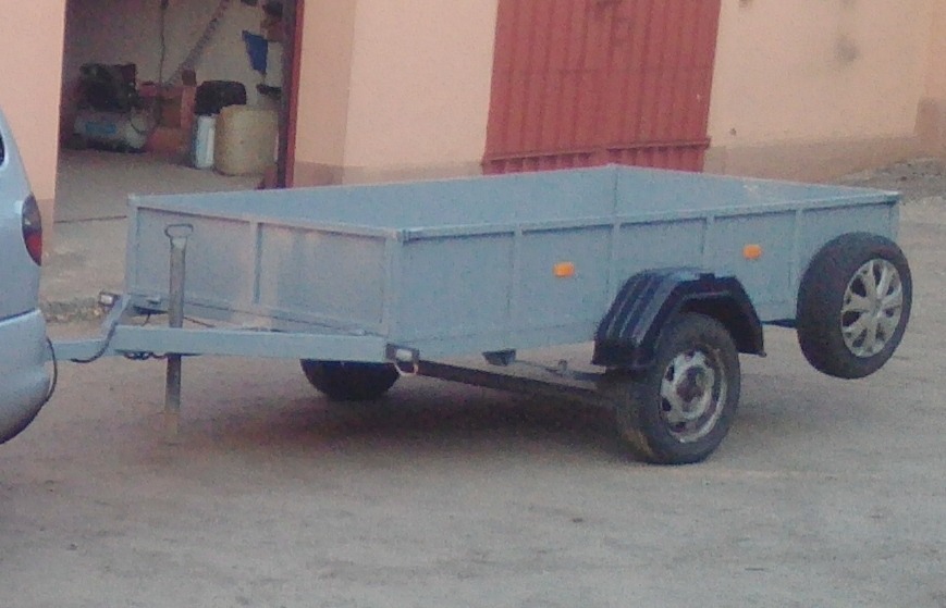 Szara przyczepka samochodowa z kołem zapasowym, zaparkowana obok samochodu na zewnątrz budynku z czerwonymi drzwiami.