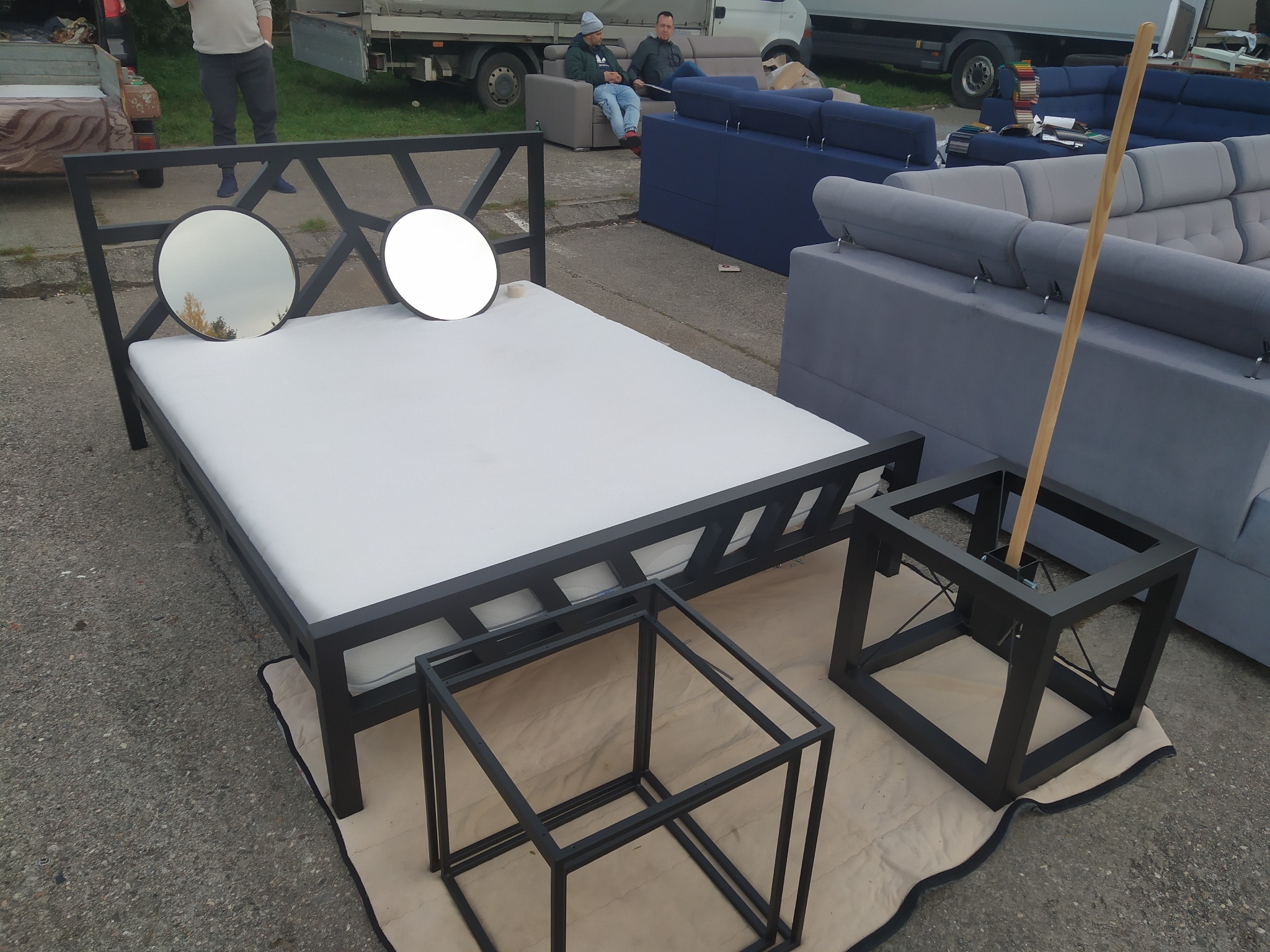 Metalowa rama łóżka z okrągłymi elementami dekoracyjnymi, ustawiona na zewnątrz obok mebli tapicerowanych i dwóch metalowych stolików o otwartej konstrukcji, z widocznymi osobami w tle.