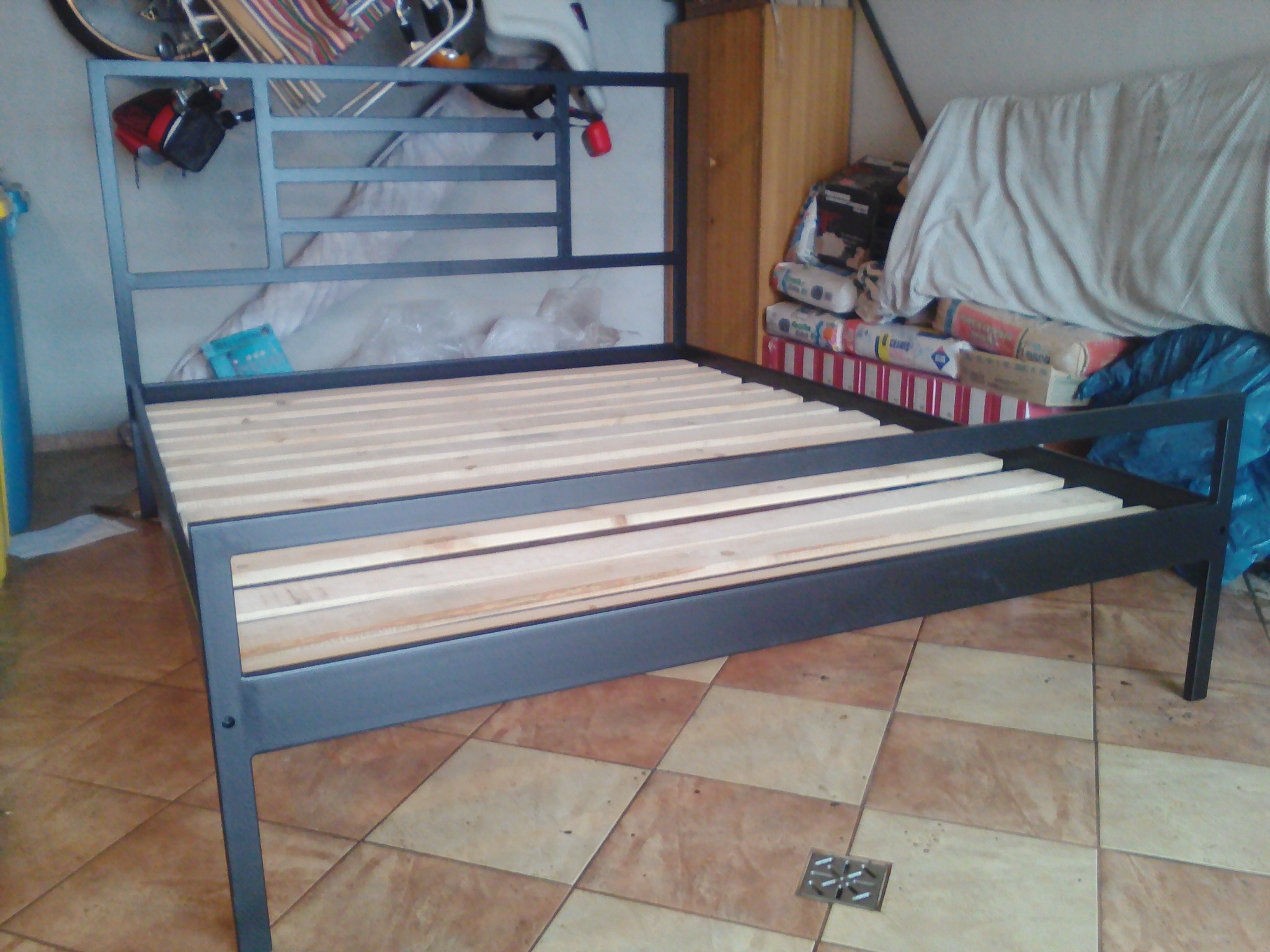 Metalowa rama łóżka z drewnianym stelażem w garażu, widoczne części roweru i materiały budowlane w tle.