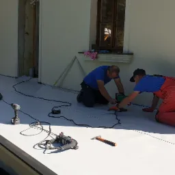 Dwóch pracowników w trakcie łączenia arkuszy białej hydroizolacji na fundamencie budynku za pomocą zgrzewarki; w tle widoczny odkurzacz przemysłowy, wiertarka i młotek.