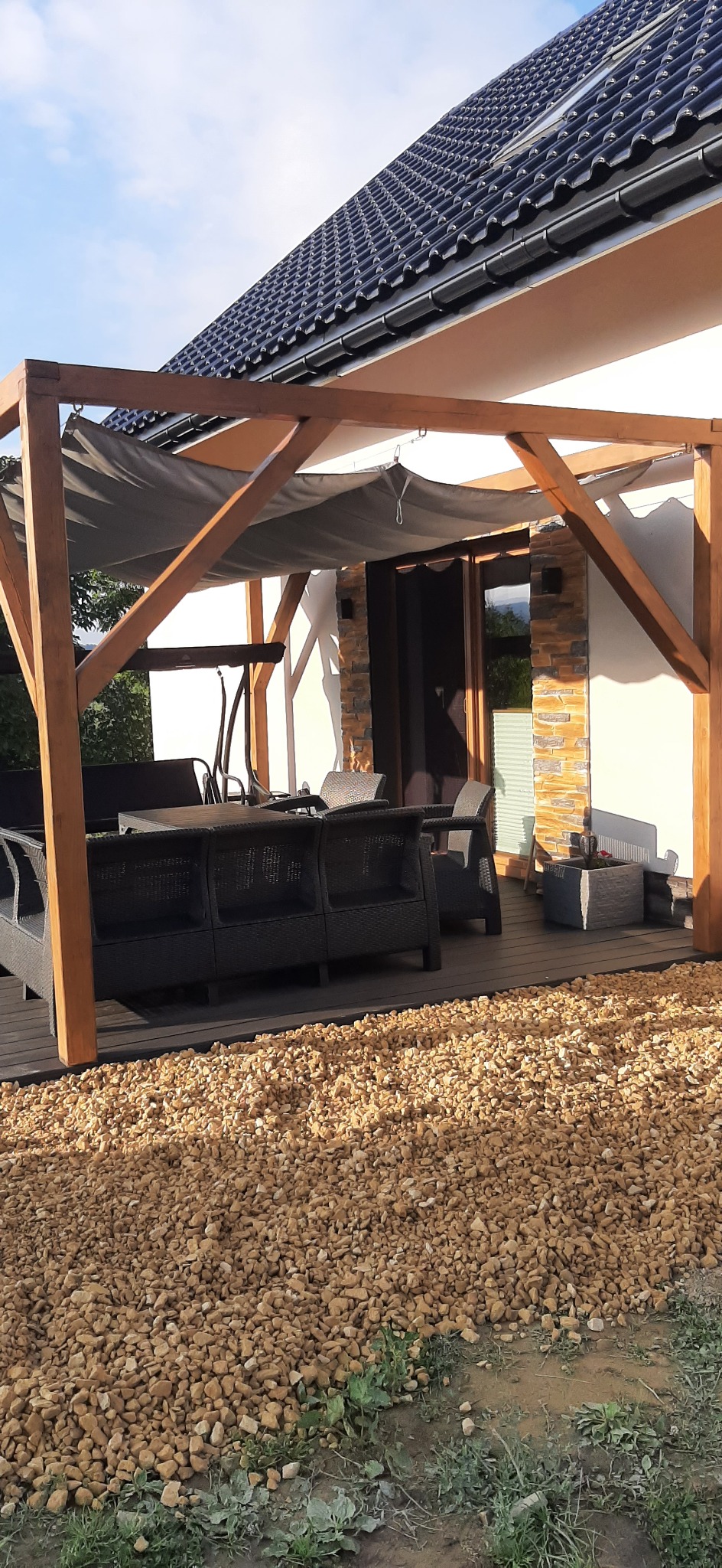 Drewniany taras z zadaszeniem w postaci beżowej tkaniny, na którym znajdują się czarne meble ogrodowe z rattanu, a obok ściany domu z kamienną okładziną.