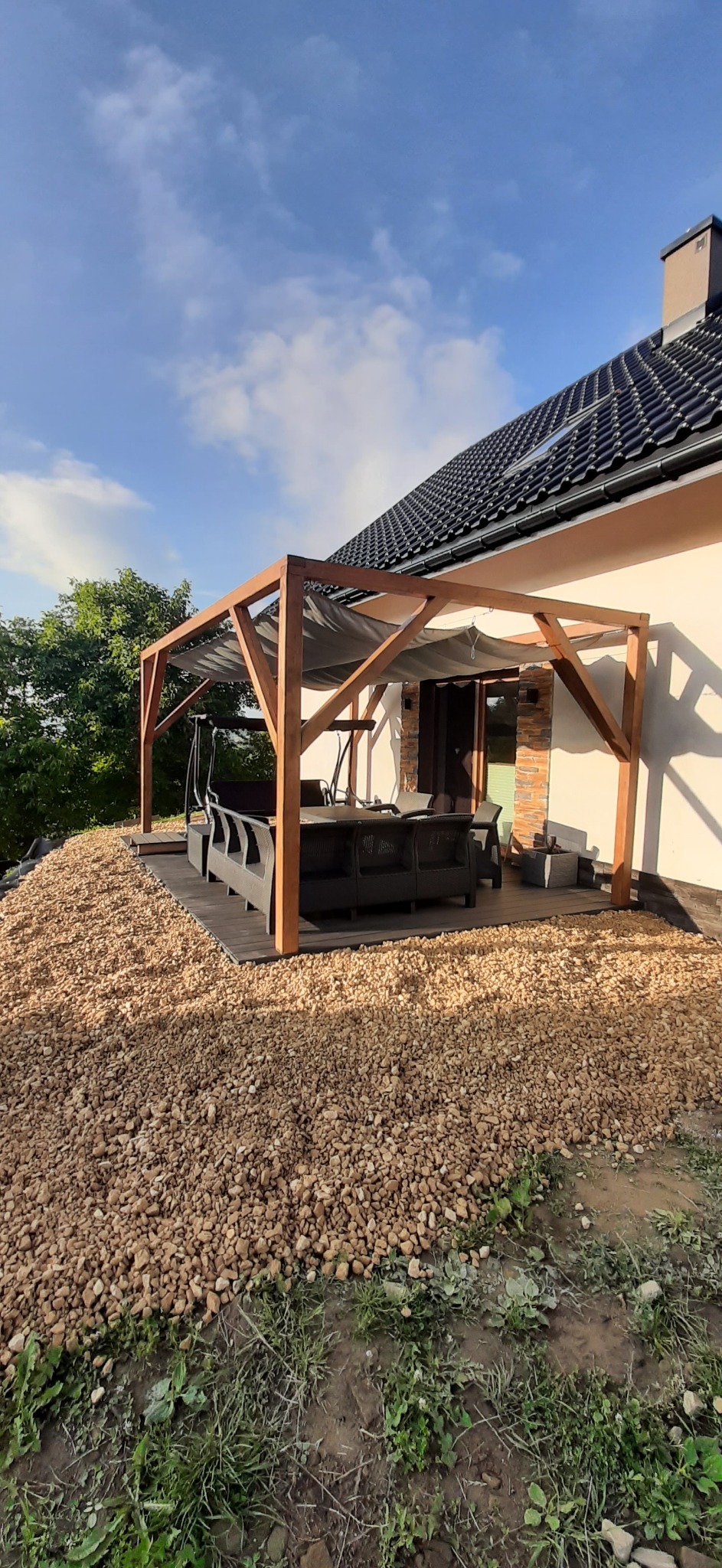 Zadaszony taras z drewnianą konstrukcją i szarą tkaniną zacieniającą, otoczony jasnymi kamieniami i zielenią, meble ogrodowe z ciemnego rattanu na deskach tarasowych.