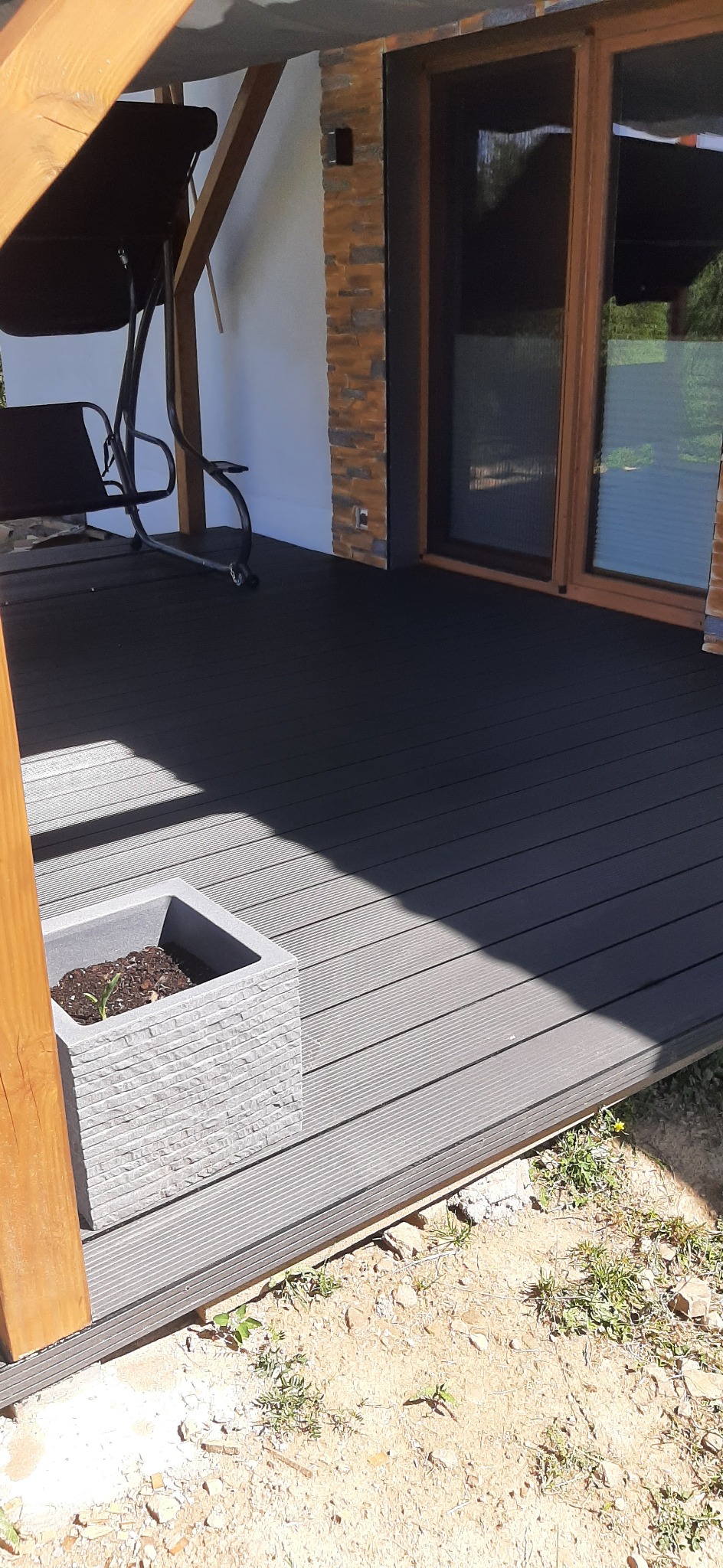 Ciemnoszary taras z desek kompozytowych, z widoczną huśtawką ogrodową, donicą z młodymi roślinami i fragmentem elewacji z kamienia dekoracyjnego.