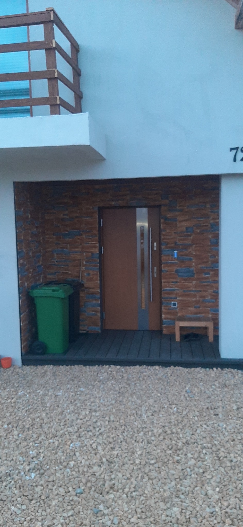 Brązowe drzwi wejściowe z pionowym, srebrnym panelem, obłożone kamieniem elewacyjnym, z widocznym numerem domu '72', zielonym koszem na śmieci i podłożem z jasnych kamyków.