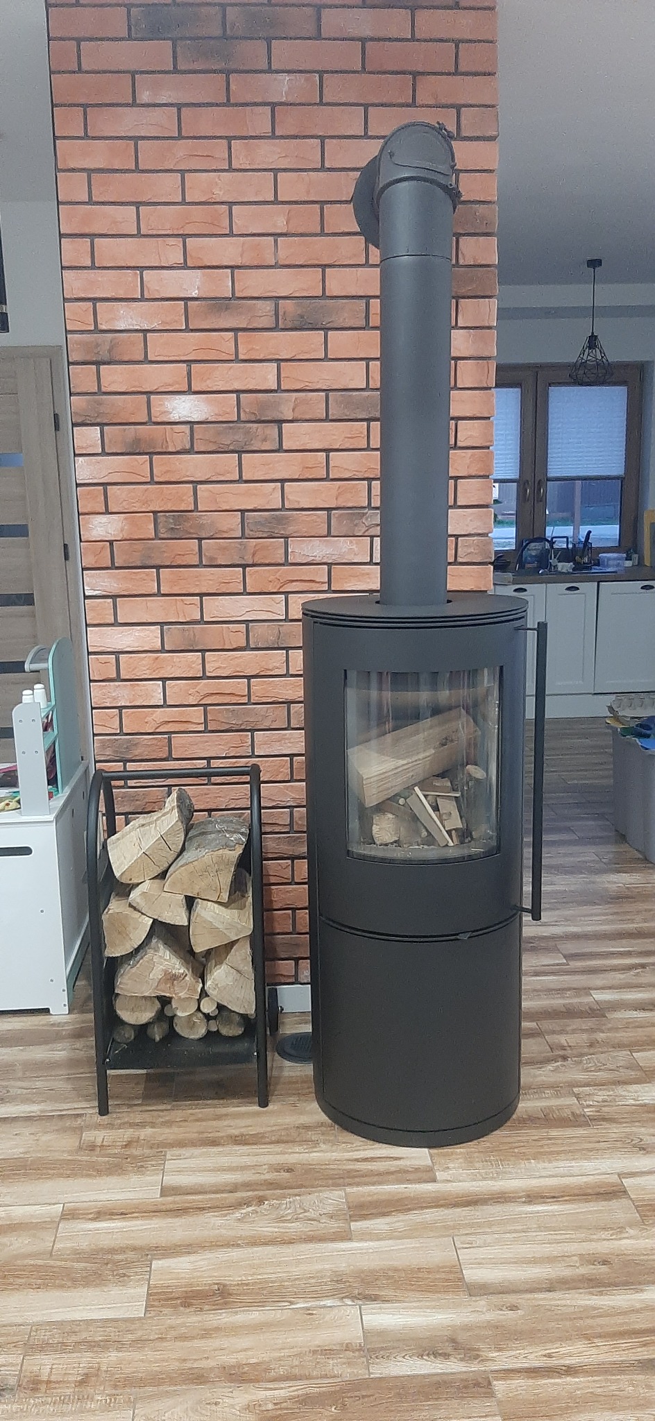 Cylindryczny, czarny piec wolnostojący z rurą odprowadzającą dym, stojący na podłodze imitującej drewno, obok metalowy stojak z ułożonym drewnem, ściana wyłożona cegłą.