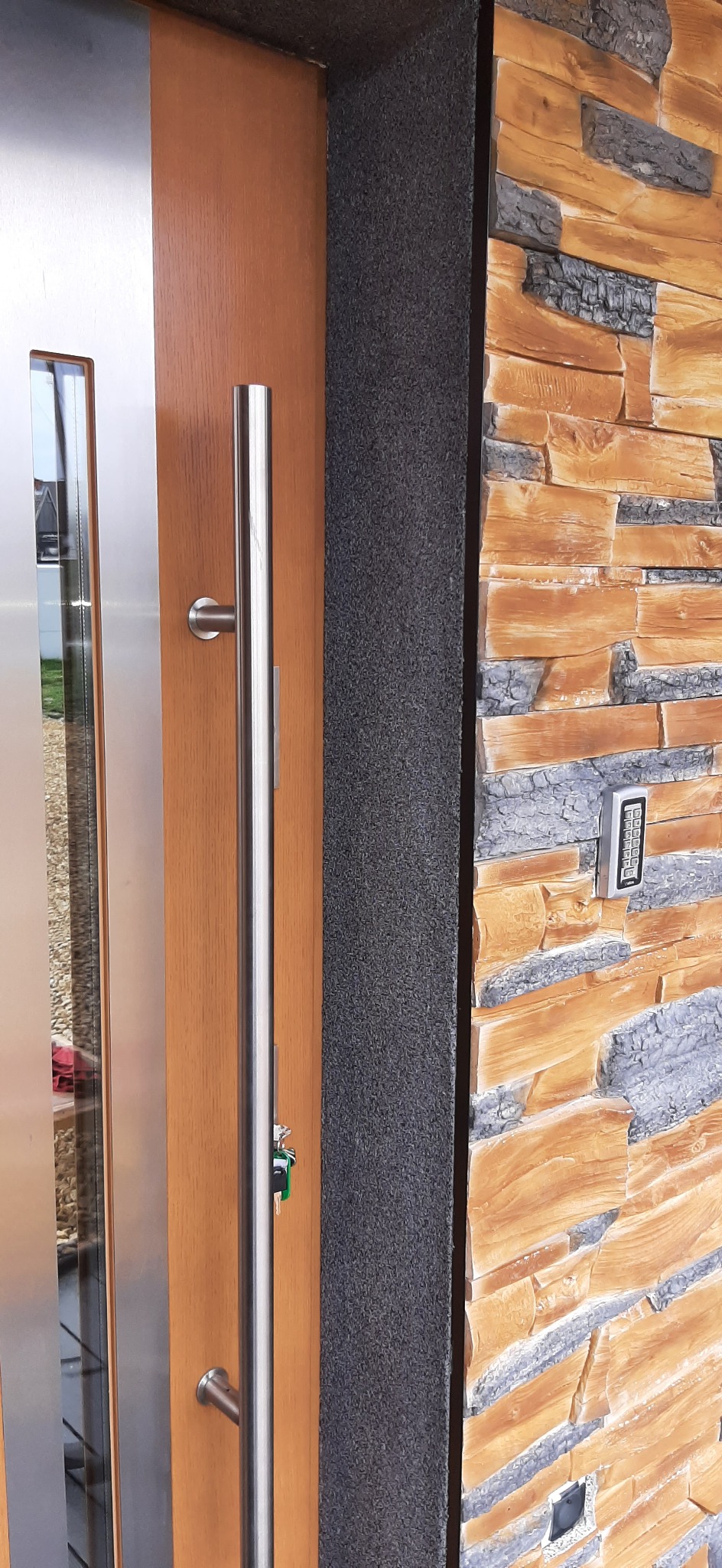 Zewnętrzne drzwi wejściowe z długim, pionowym uchwytem ze stali nierdzewnej, obok ściana wykończona dekoracyjnym kamieniem imitującym drewno i szarym, chropowatym tynkiem.