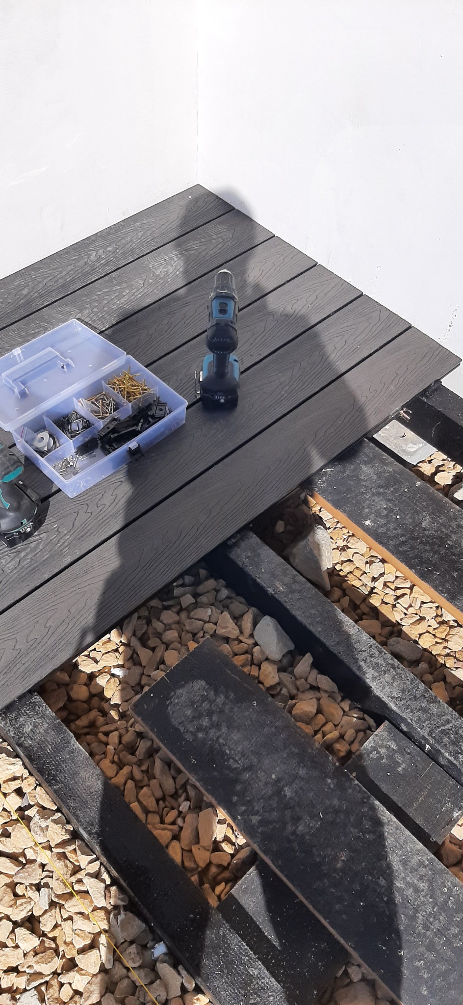 Montaż tarasu z desek kompozytowych: widoczny fragment konstrukcji, skrzynka z elementami montażowymi, wkrętarka akumulatorowa i podsypka z kamieni.