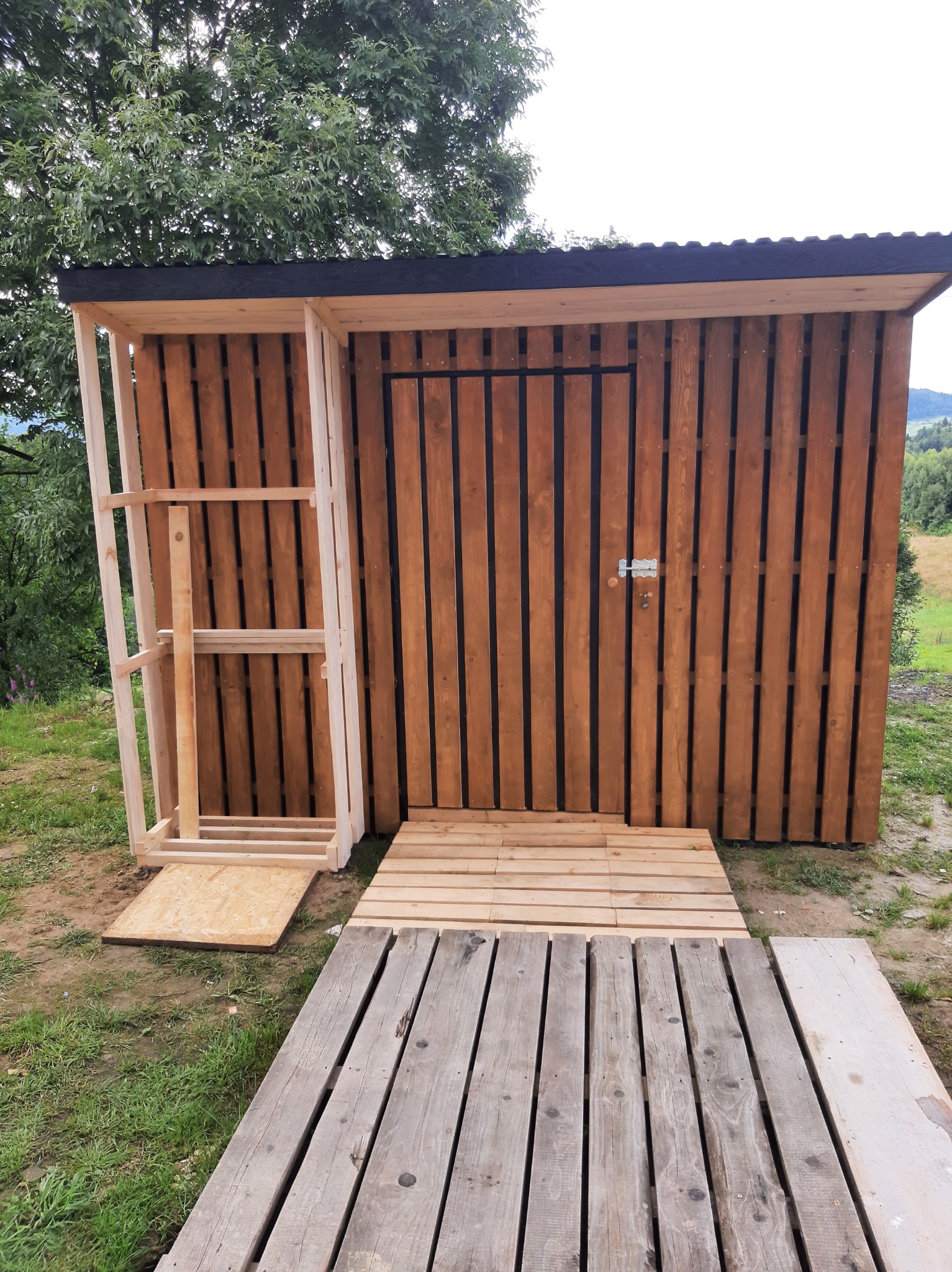 Drewniana konstrukcja szopy ogrodowej w trakcie budowy, z widocznym szkieletem i częściowo ukończoną ścianą z desek, położona na trawiastym terenie z elementami drewnianej podłogi z palet.