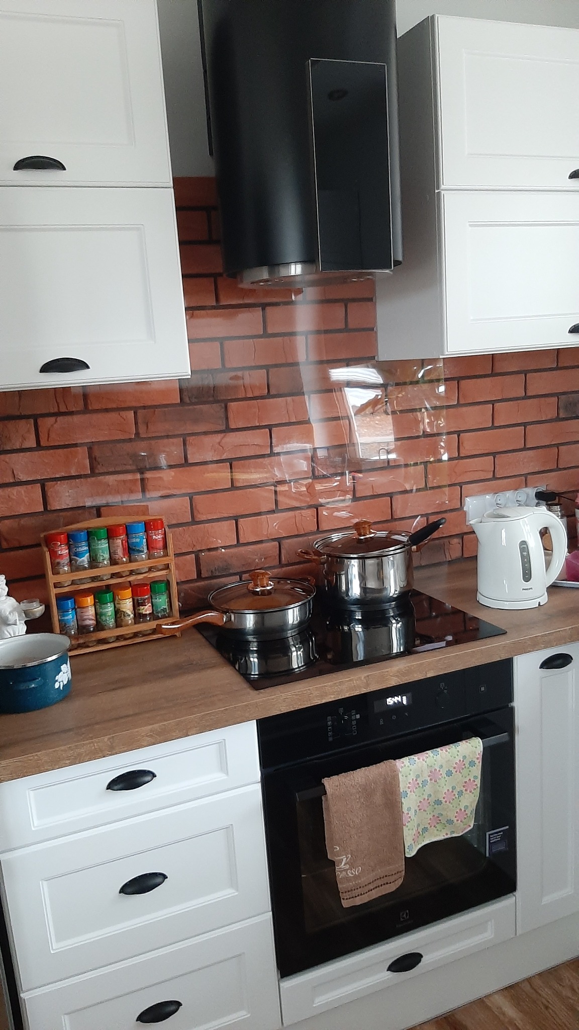 Nowoczesna kuchnia z białymi szafkami z czarnymi uchwytami, ceglaną ścianą nad blatem i czarnym okapem. Na indukcyjnej płycie grzewczej stoją dwa garnki, obok czajnik elektryczny, a na blacie...