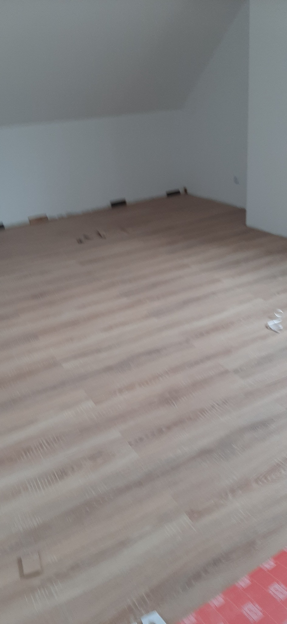 Ułożona podłoga z paneli w jasnym odcieniu drewna, widoczne niedokończone fragmenty przy ścianie i listwy przypodłogowe jeszcze nie zamontowane, surowy stan pomieszczenia.