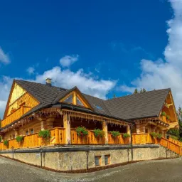 Gotowy dom z bali z ciemnym dachem, oświetleniem dekoracyjnym i balkonem, z kwiatami w skrzynkach, na kamiennej podmurówce, pod błękitnym niebem.