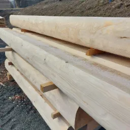 Stos jasnych, obrobionych bali drewnianych, ułożonych na przekładkach, z widocznym rysunkiem słojów, na tle żwirowego podłoża i zalesionego wzgórza.