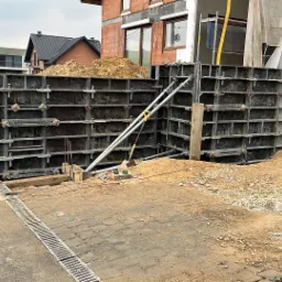 Szare szalunki stalowe przygotowane do zalania betonem, tworzące formę dla przyszłej ściany oporowej, widoczne podpory i wykop, w tle budynek w trakcie budowy.