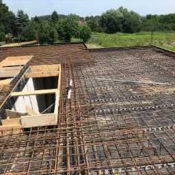 Zbrojenie płyty fundamentowej z widocznymi otworami na instalacje, szalunki drewniane i elementy kanalizacyjne, w tle zieleń drzew i krzew&oacute;w.