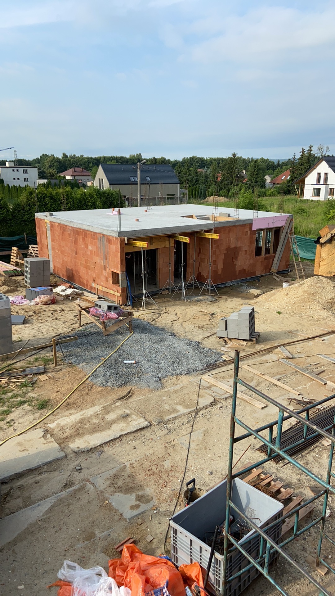 Wznoszenie ścian parterowego budynku z czerwonej cegły, widoczne szalunki i podpory stropu, teren budowy z gruzem i materiałami.