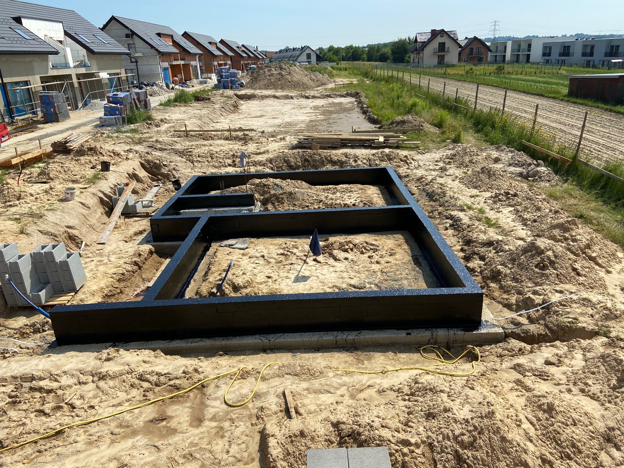Przygotowany szalunek fundamentów pomalowany na czarno, widoczne bloczki betonowe i wykop, w tle zabudowa jednorodzinna w trakcie budowy.