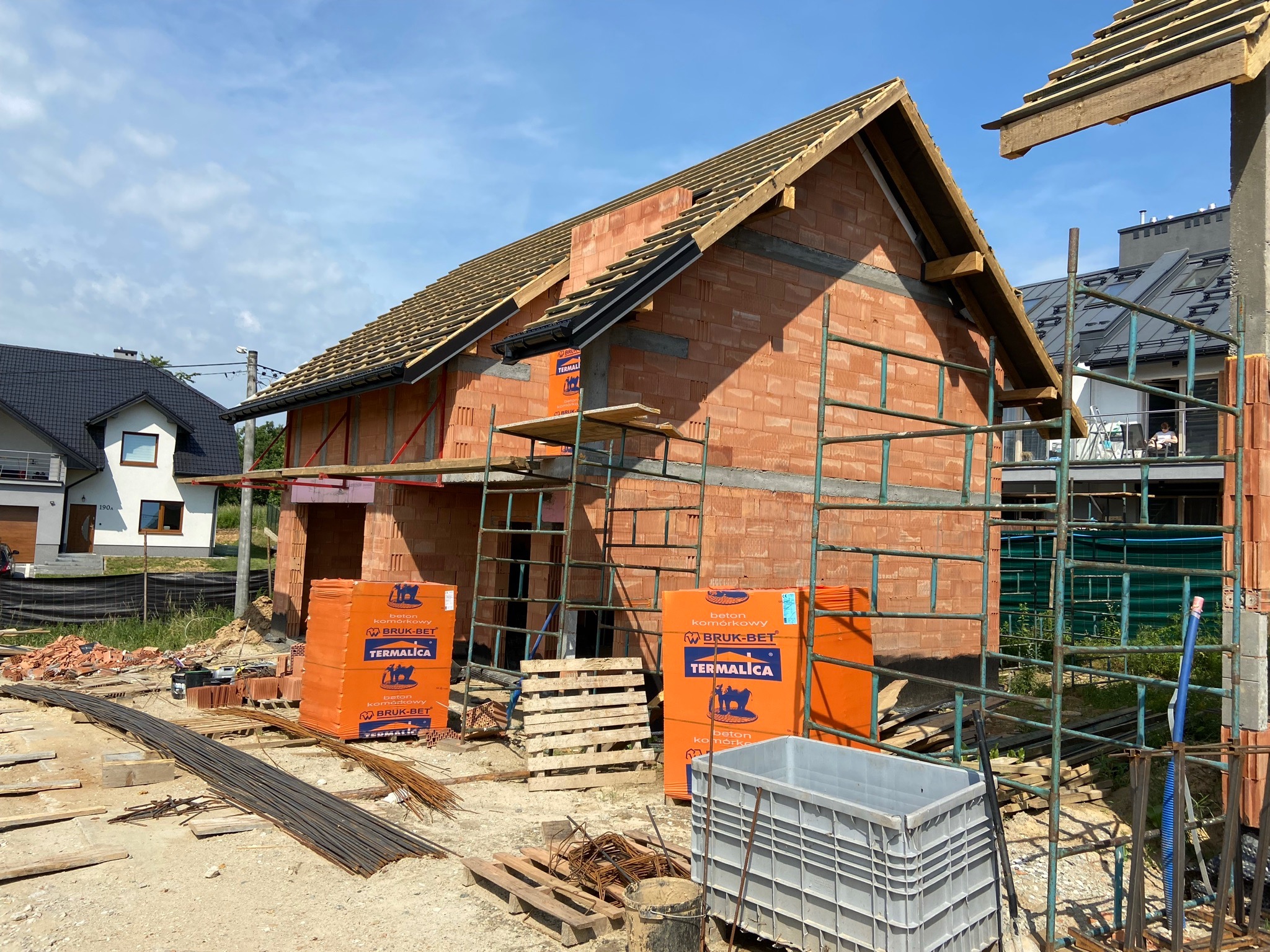 Wznoszenie ścian z bloczków komórkowych Termalica na budowie domu jednorodzinnego; widoczne rusztowania, palety z materiałami i zbrojenie.