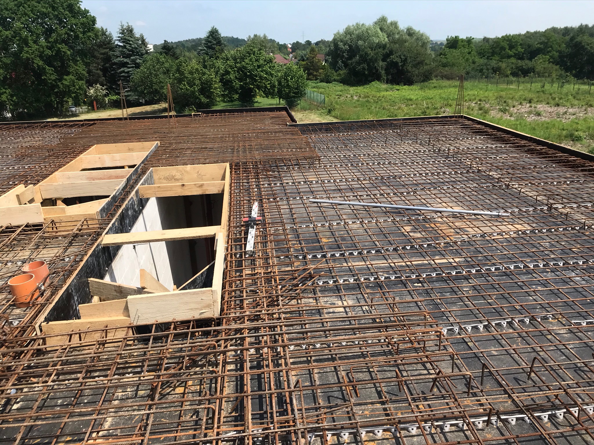 Zbrojenie płyty fundamentowej z widocznymi otworami na instalacje, szalunki drewniane i elementy kanalizacyjne, w tle zieleń drzew i krzewów.