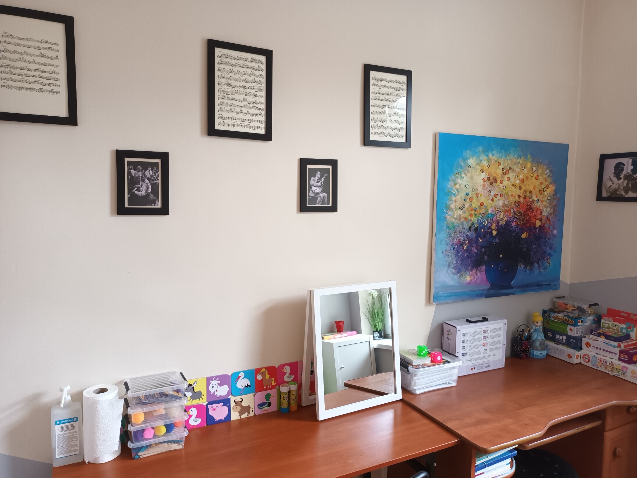 Pokój z biurkiem, na którym znajdują się pomoce edukacyjne, lustro i obraz. Na ścianie wiszą oprawione nuty i zdjęcia.