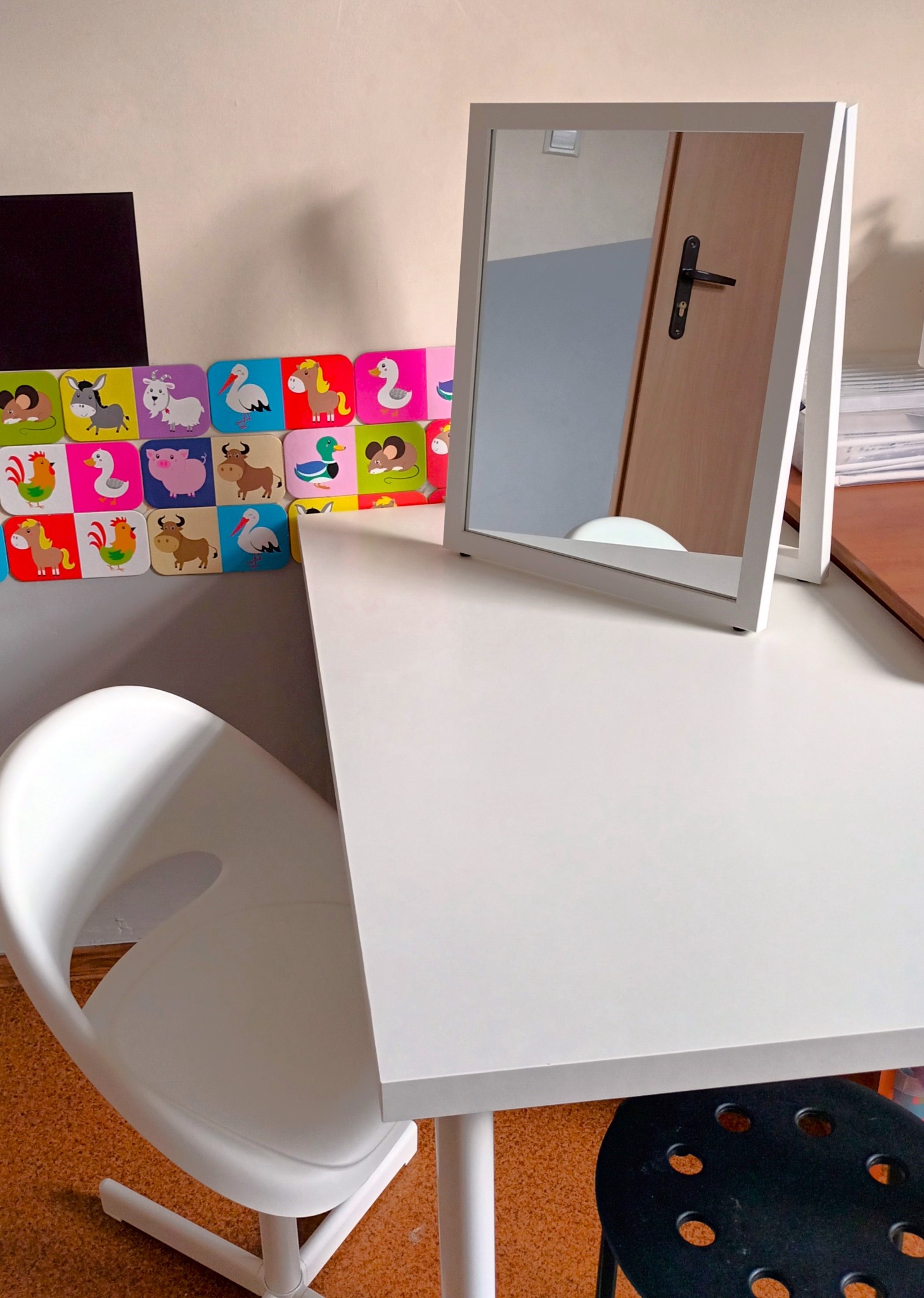 Białe biurko z lustrem ramkowym i krzesłem, ozdobione kolorowymi kartami z wizerunkami zwierząt, tworząc przestrzeń do terapii dziecięcej.