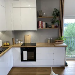 Biała kuchnia na wymiar z drewnianym blatem, płyta indukcyjna, piekarnik Samsung i drewniane półki z książkami i roślinami, z widokiem na balkon i ogród, na pierwszym planie biały pies rasy West...