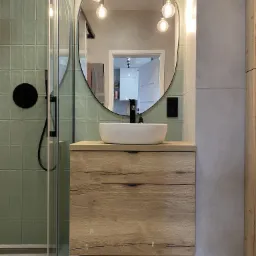 Nowoczesna łazienka z szafką pod umywalkę z drewnopodobnym frontem, owalnym lustrem i wiszącymi żarówkami edisona, widoczna kabina prysznicowa z czarnym panelem natryskowym.