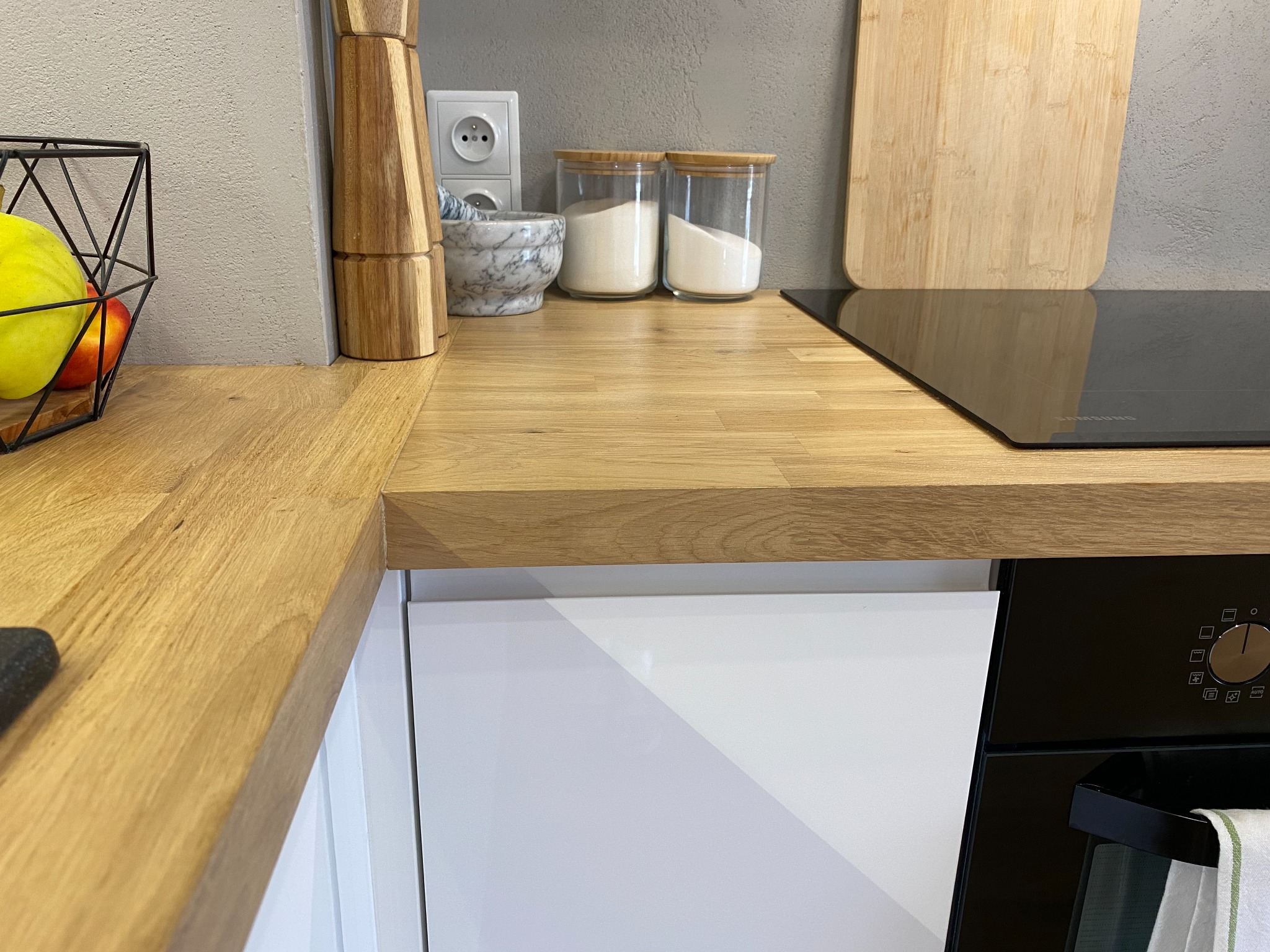 Fragment kuchni z drewnianym blatem, na którym stoją przyprawy w szklanych pojemnikach, moździerz i deska do krojenia, obok płyty indukcyjnej i białych szafek.