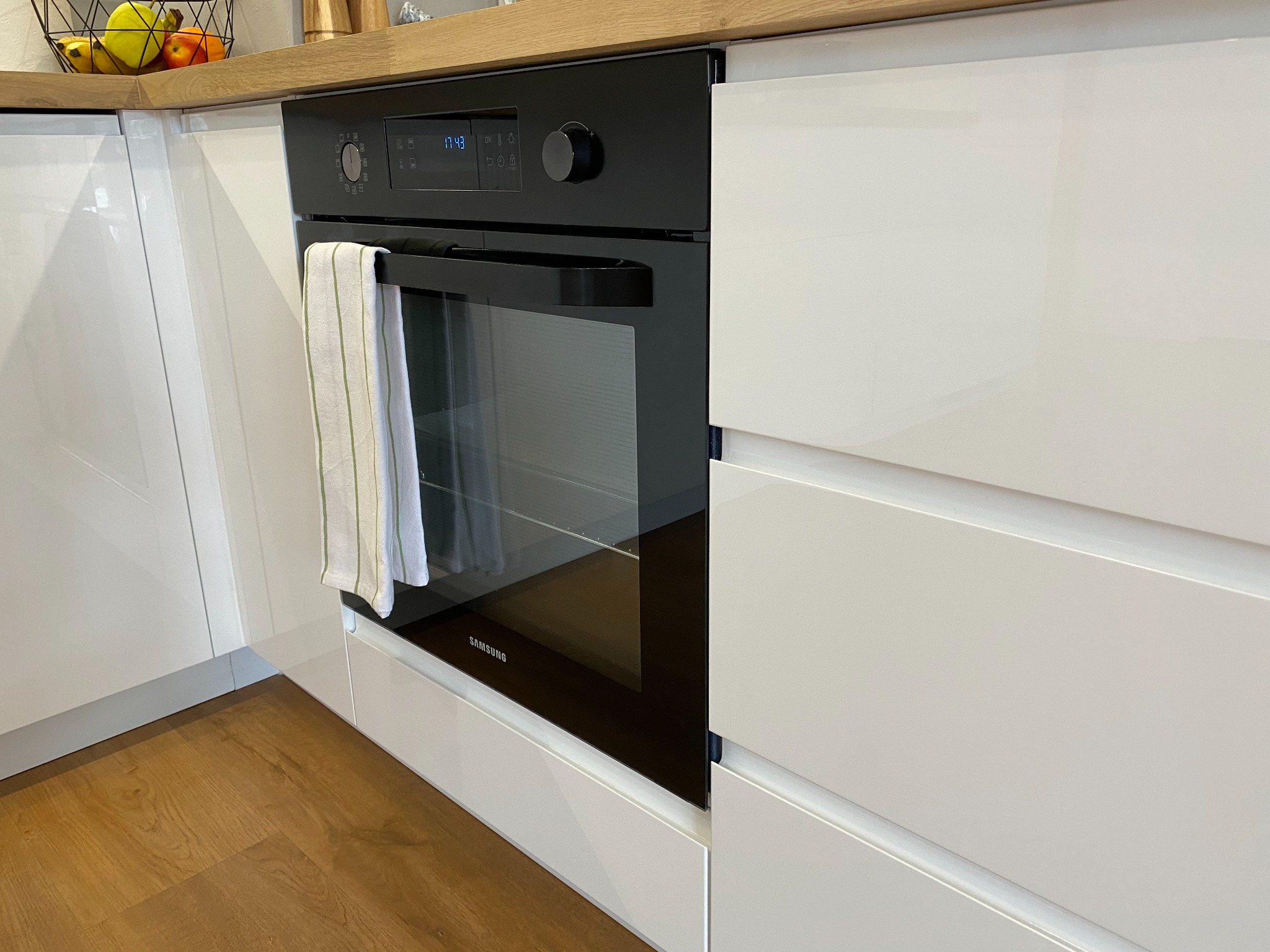Czarny piekarnik Samsung w zabudowie z białych, lakierowanych frontów kuchennych z drewnianym blatem, widoczny fragment podłogi z paneli.