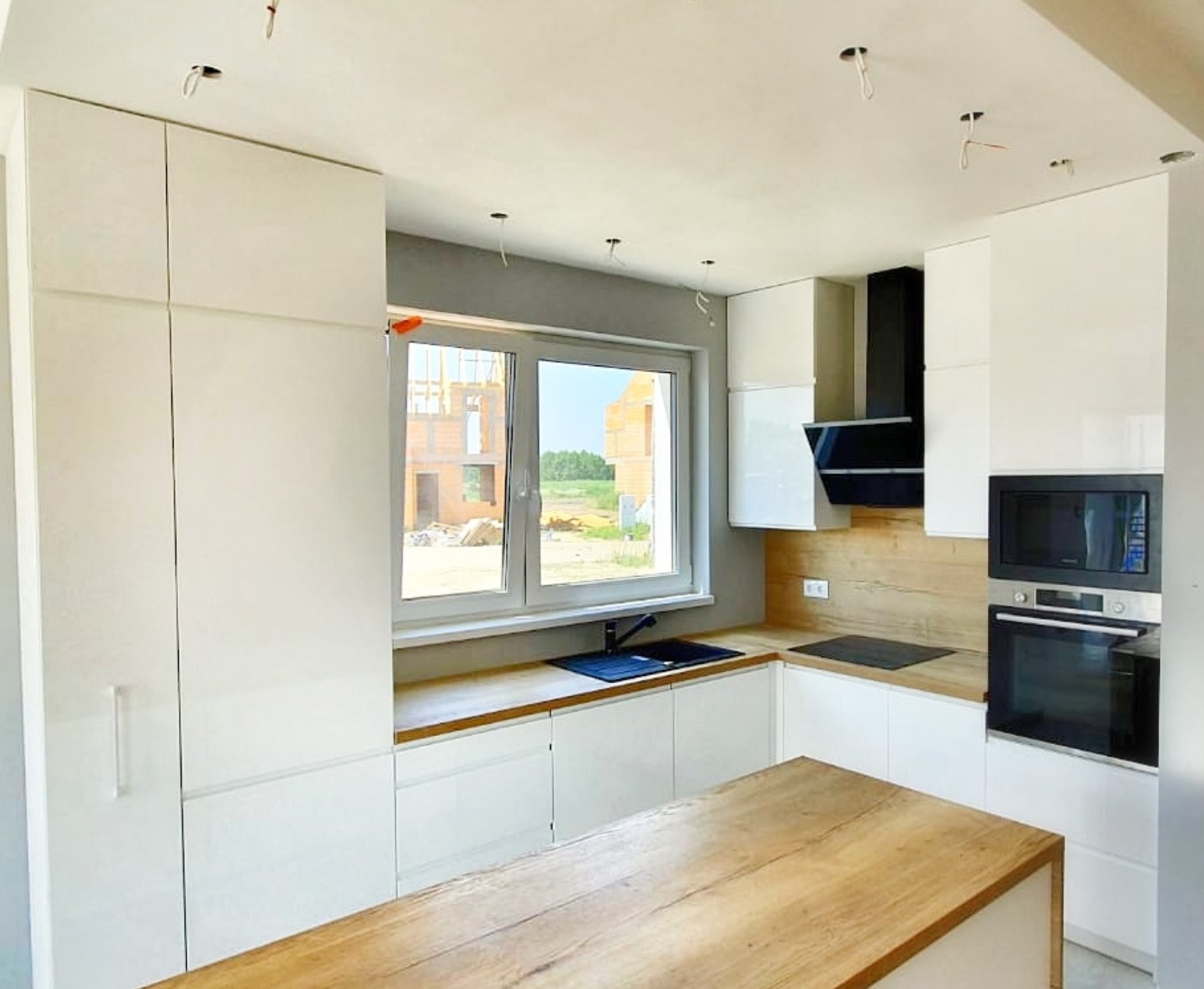 Nowoczesna kuchnia z białymi, lakierowanymi frontami, drewnianym blatem i wyspą kuchenną w trakcie montażu, widoczne niedokończone podłączenia elektryczne.