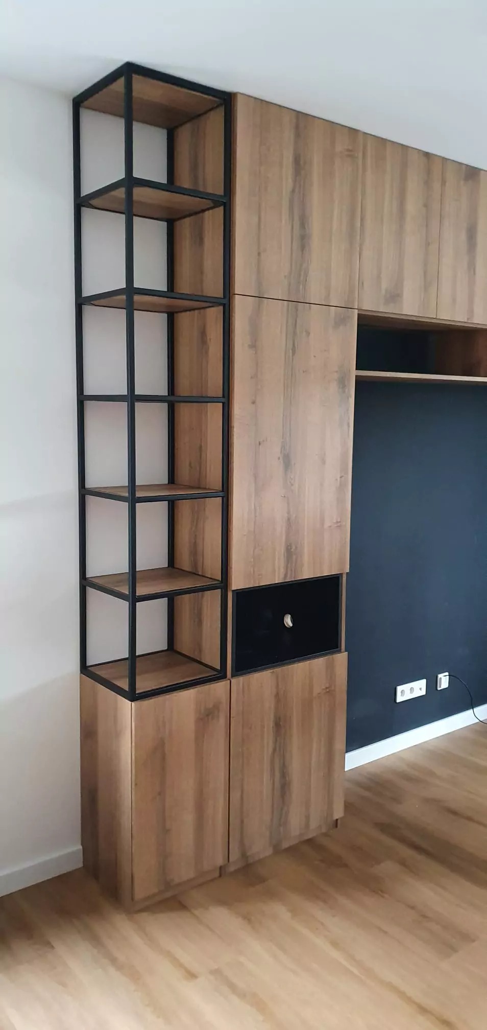 Nowoczesna zabudowa meblowa z drewnianymi frontami i czarnym regałem z metalu, wnęka na sprzęt RTV na tle granatowej ściany.