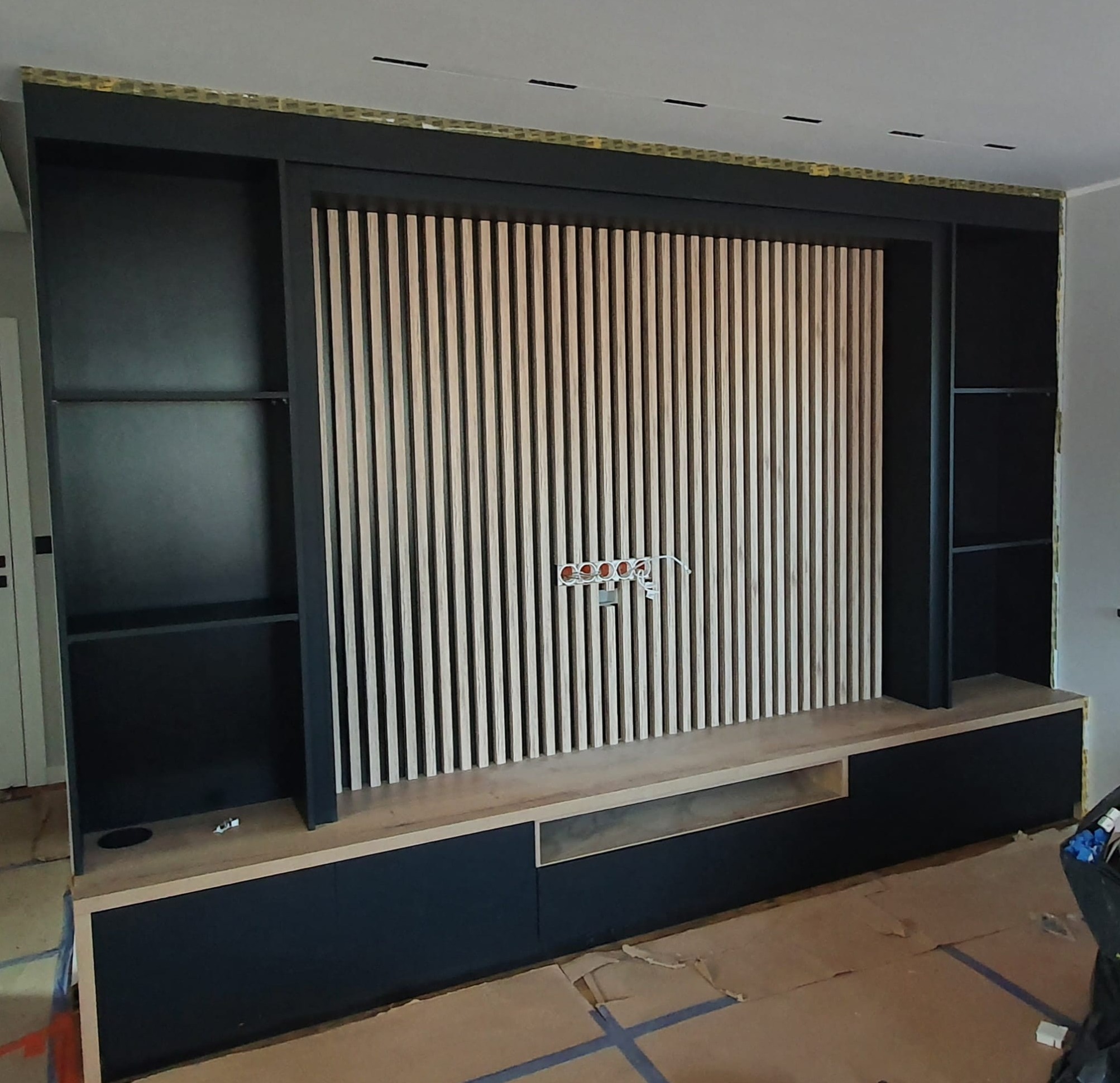 Częściowo zmontowana, czarno-beżowa zabudowa ściany z drewnianymi lamelami i otwartymi półkami, widoczne gniazdka elektryczne i karton na podłodze.