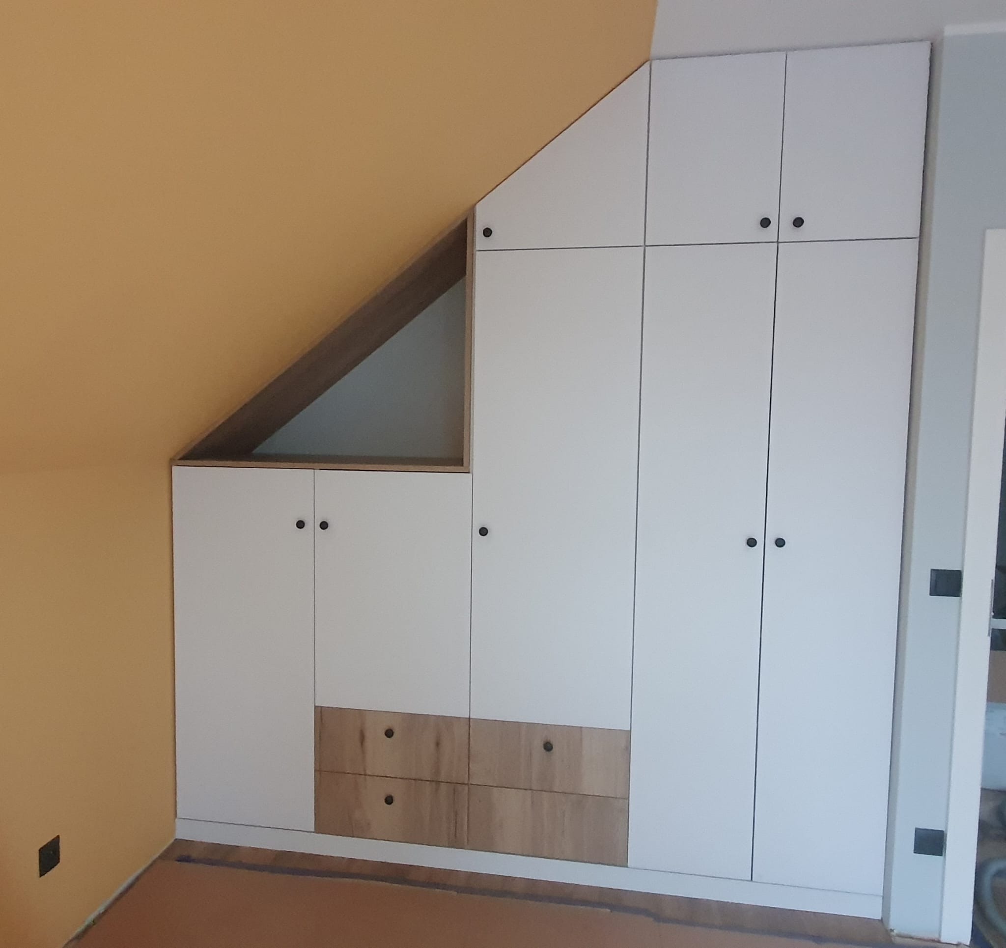 Biała zabudowa szafy z drewnianymi akcentami w skosie poddasza, z ciemnymi okrągłymi uchwytami.