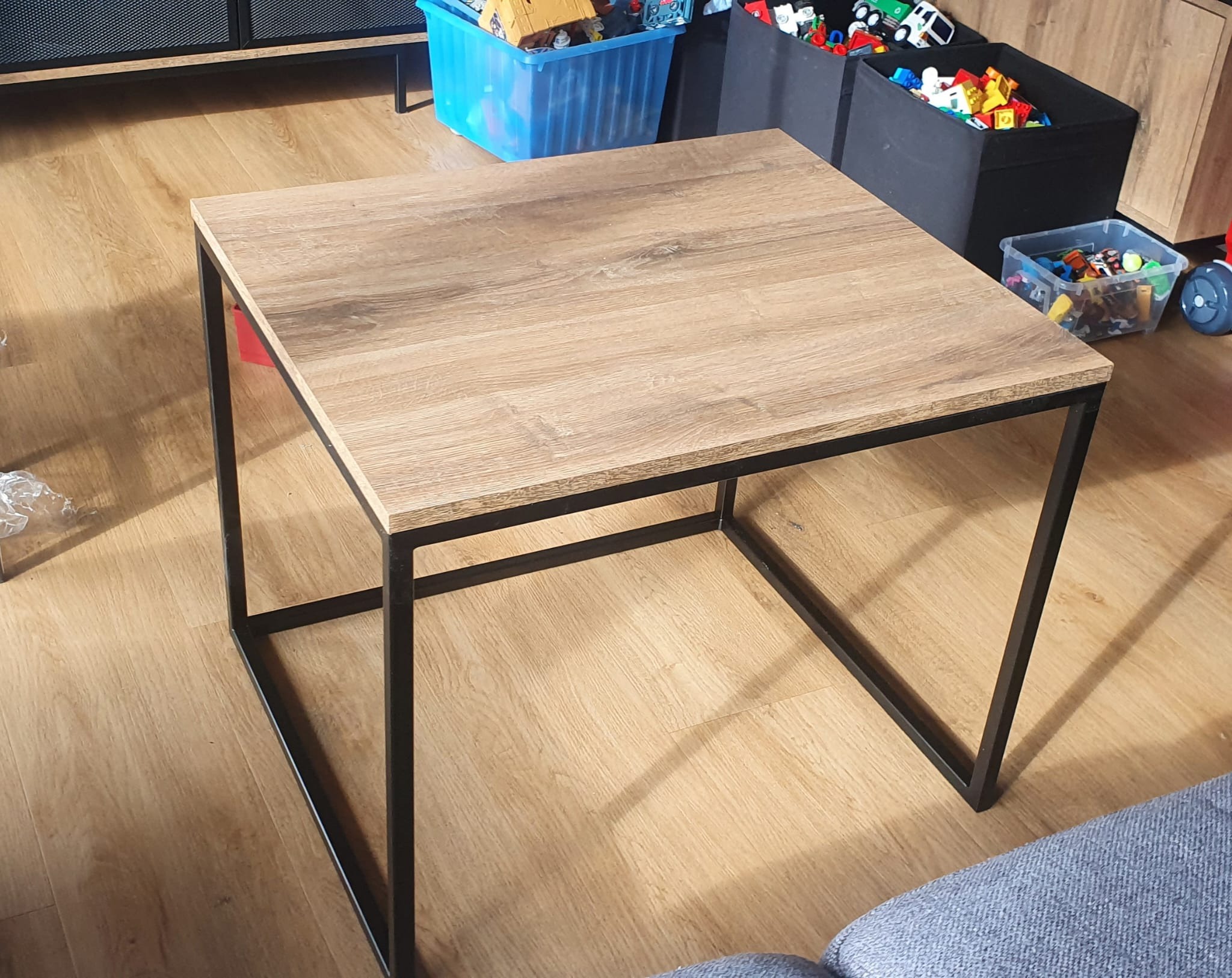 Kwadratowy stolik kawowy z blatem imitującym jasne drewno i czarną, metalową podstawą na tle podłogi w odcieniach brązu i pojemników z zabawkami.