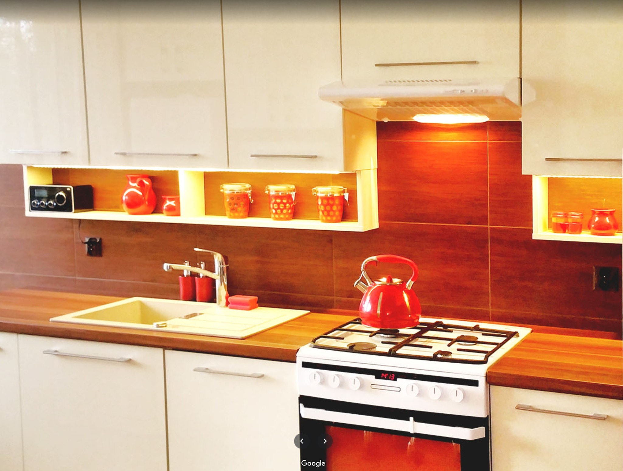 Jasna kuchnia z drewnianym blatem, płytkami w odcieniach brązu, białymi szafkami z poziomymi uchwytami i czerwonym czajnikiem na kuchence.