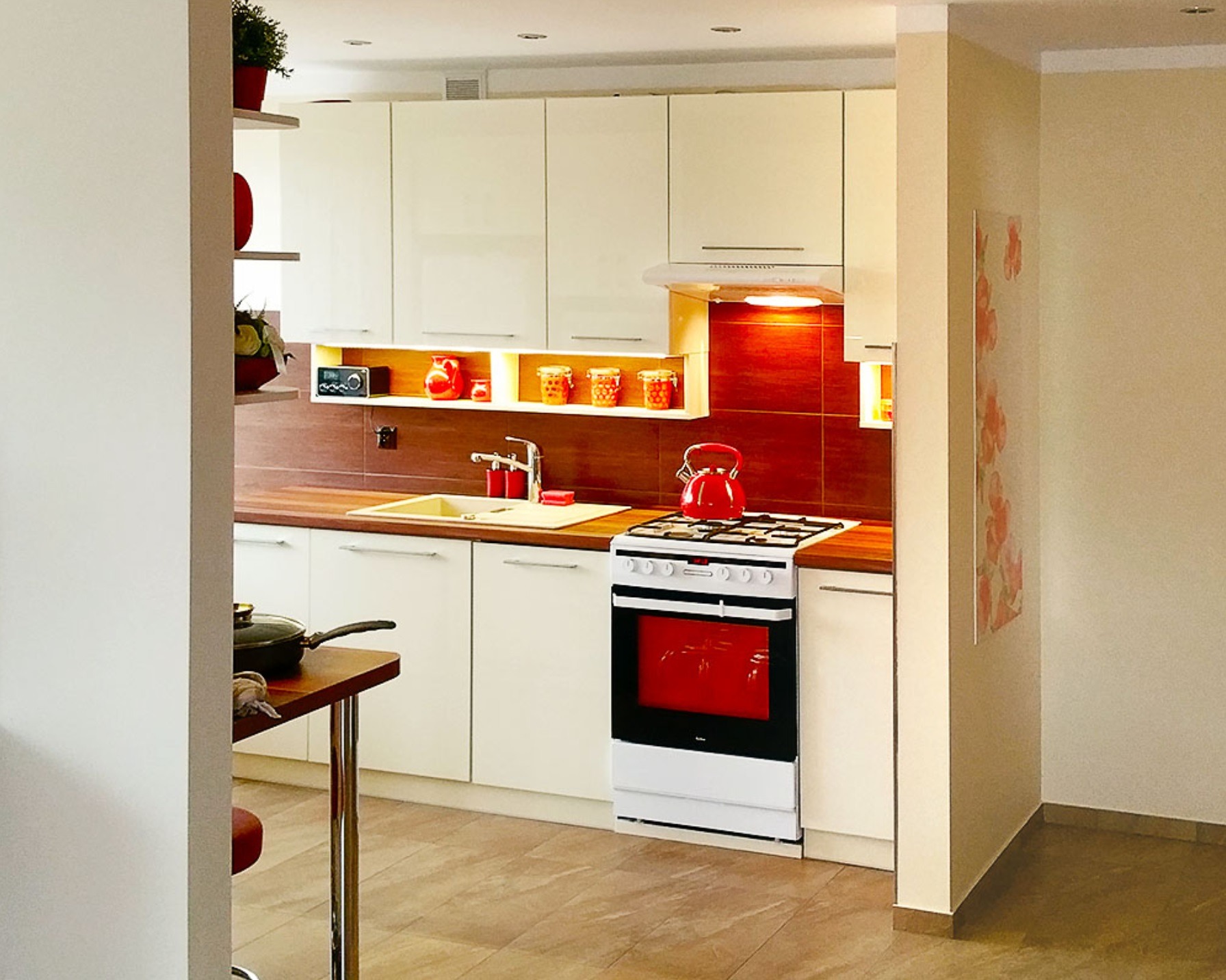 Nowoczesna kuchnia z białymi szafkami, drewnianym blatem i czerwoną ścianą nad blatem, widoczna przez wejście do pomieszczenia, z garnkiem na stole w tle.