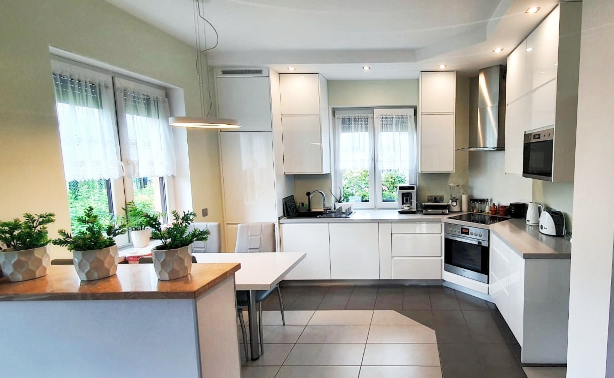 Jasna kuchnia z białymi, błyszczącymi frontami szafek, szarym blatem i podłogą, z widocznym stołem i dekoracyjnymi roślinami w doniczkach na blacie oddzielającym kuchnię od jadalni.