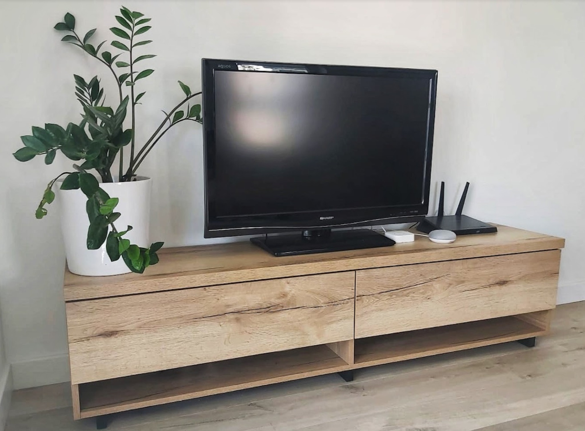 Drewniana szafka RTV z dwoma szufladami i otwartą półką, na której stoi telewizor, głośnik inteligentny i router, obok doniczka z zieloną rośliną, całość w jasnym, minimalistycznym wnętrzu.