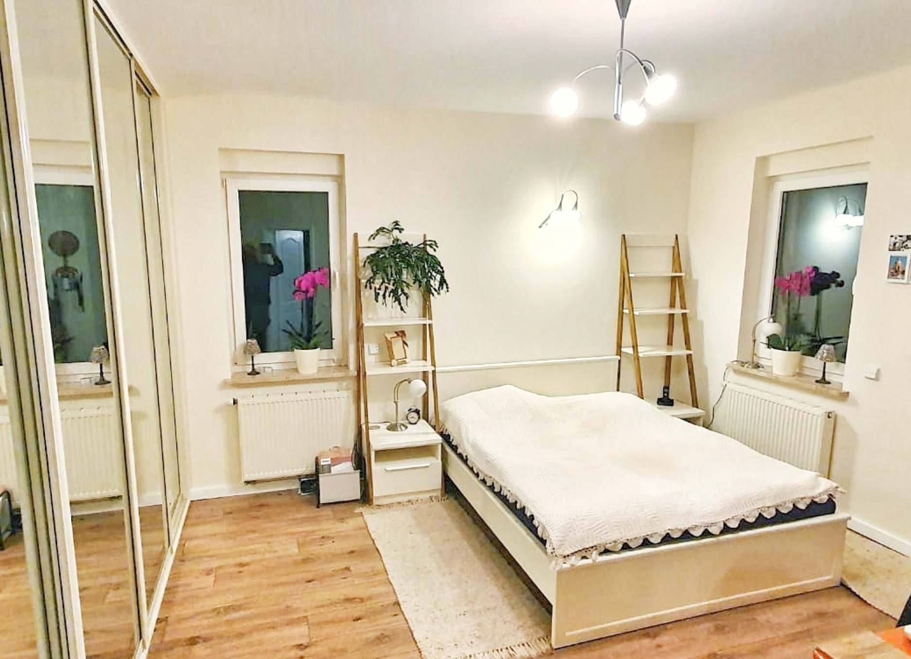 Jasna sypialnia w Poznaniu z drewnianą podłogą, białym łóżkiem z ozdobnym wykończeniem, szafą z lustrzanymi drzwiami przesuwnymi i drewnianymi regałami po bokach łóżka.