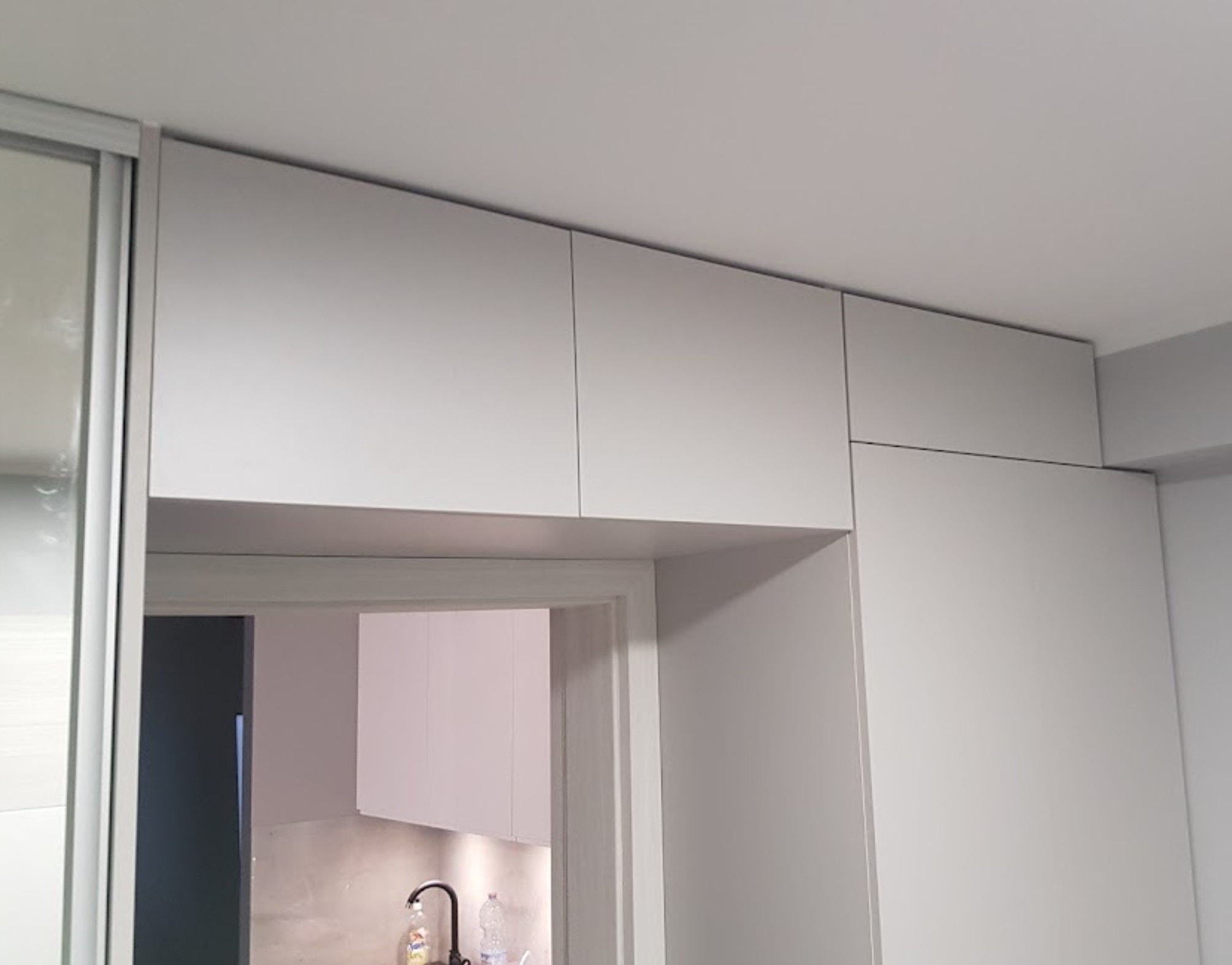 Szara zabudowa meblowa pod sufitem, z szafą przesuwną po lewej i widokiem na kuchnię z czarną baterią, widziana z perspektywy dolnej.